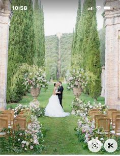 a bride and groom standing in front of an archway with flowers on the side, surrounded by greenery