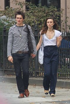 a young man and woman walking down the street holding hands while wearing overalls, striped shirt and black pants