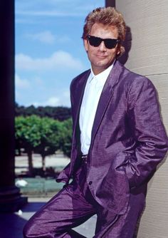 a man in a purple suit and sunglasses leaning against a pillar with his hands on his hips