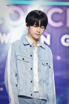 a young man with black hair wearing a denim jacket and white shirt, standing in front of a blue background