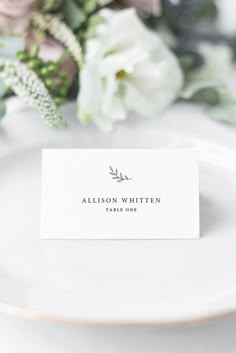 a place card sitting on top of a white plate with flowers in the back ground