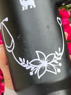 a hand holding a black coffee mug with white designs on it and pink flowers in the background