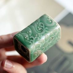 a hand holding a green and white object in it's left hand, with an ornate design on the top