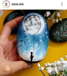 someone is holding up a painted rock with a boy standing on the moon and birds flying over it