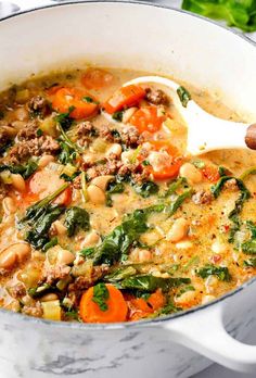 a white pot filled with meat and vegetables on top of a counter next to a wooden spoon