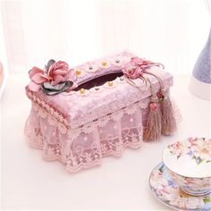 a pink lace box with flowers and tassels on it next to tea cups