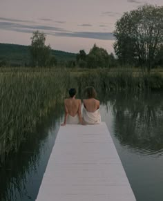 two women sitting on a dock looking out at the water and grass in the background