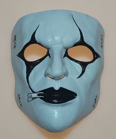 a blue mask with orange eyes and nose piercings on the face is shown in front of a white wall