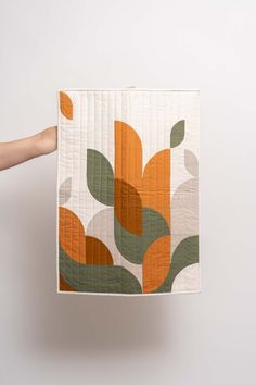 a person holding up a quilt with orange and green leaves on it, in front of a white wall