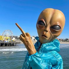 an alien man pointing at the camera with a ferris wheel in the back ground behind him