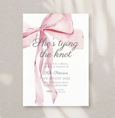 a pink ribbon tied around the back of a white card with text that reads, she's tying the knot