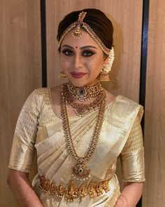 a woman in a white and gold sari with jewelry on her neck, standing next to a wooden wall