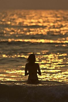 a woman standing in the ocean at sunset with her back to the camera, facing the water