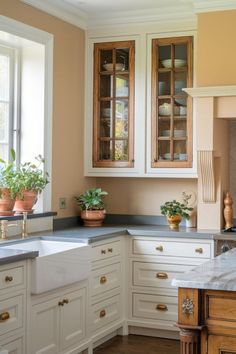 Farmhouse white kitchen with glass cabinet doors, brass pulls, gray counter tops, and potted herbs in the kitchen window White Kitchen With Brass Hardware, Kitchen With Brass Hardware, Gray Counter Tops, Potted Herbs