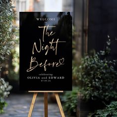 a black and gold welcome sign sitting on top of a wooden easel in front of some plants