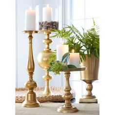 three candles are sitting in gold vases with ferns and pine cones on the table