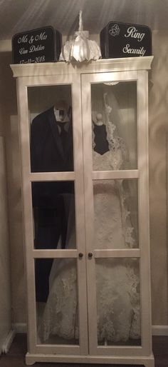 a wedding dress and tuxedo on display in a white cabinet with glass doors