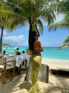 a woman in a yellow skirt standing on the beach next to palm trees and water