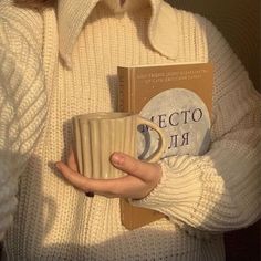 a woman holding a book and coffee mug in her hands while wearing a white sweater