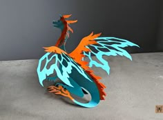 a paper sculpture of a dragon with orange and blue wings sitting on cement ground next to gray wall