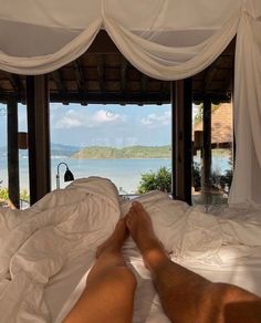 a man laying in bed with his feet up on the pillow and looking out at the ocean