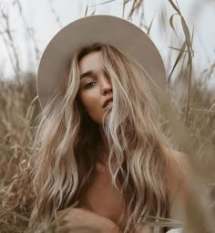 a woman with long blonde hair wearing a white hat and sitting in tall grass looking at the camera