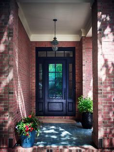 a black door and some plants in front of it on a brick building with light coming through the window