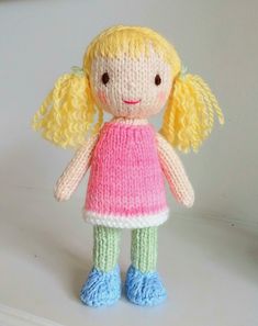 a small knitted doll with blonde hair and green leggings stands on a white surface