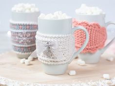 three coffee mugs decorated with crochet are sitting on a wooden tray next to marshmallows