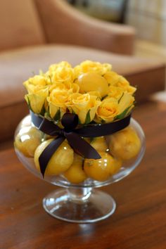 a glass bowl filled with yellow roses and lemons on top of a wooden table