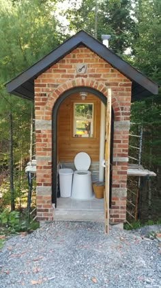 a toilet in a small brick building surrounded by trees and graveled area with steps leading up to it