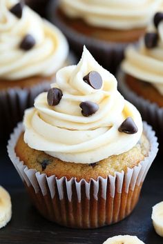 cupcakes with white frosting and chocolate chips are on a black surface surrounded by bananas