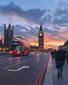 a red double decker bus driving down a street next to a tall tower with a clock on it's side