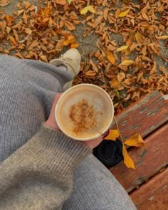 a person sitting on a bench holding a cup of coffee in their hand with leaves around them