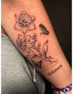 Black and white tattoo of two daffodils intertwined with smaller flowers on the forearm. There is a small butterfly to the right of the flowers, and Roman numerals underneath.  Everything has light grey shading and white highlights. Daffodil Half Sleeve Tattoo, Narcissus Flower Tattoo With Butterfly, Bird Flower Tattoo Design, Tattoo With Lily Flowers, Flower Tattoo With Roman Numerals, Daffodil Name Tattoo, Daffodil With Butterfly Tattoo, Butterfly Daffodil Tattoo, Flower And Roman Numeral Tattoo