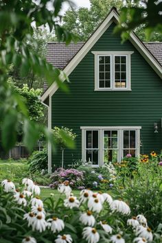 a green house with white windows and lots of flowers in the front yard, surrounded by greenery