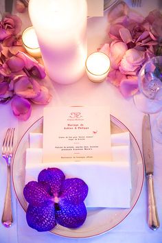 the table is set with purple flowers, candles and napkins for dinner guests to eat