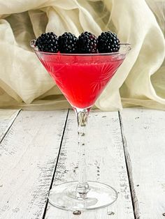 a martini glass filled with blackberries on top of a white wooden table next to a blanket