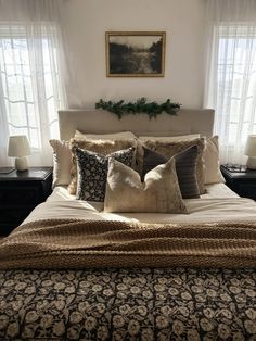 a bed with many pillows on it in front of two windows and a painting above the headboard