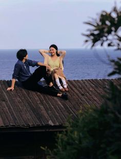 two people sitting on top of a wooden roof next to the ocean and one person standing up