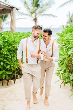 two men in white shirts and tan suspenders are walking down the beach with palm trees