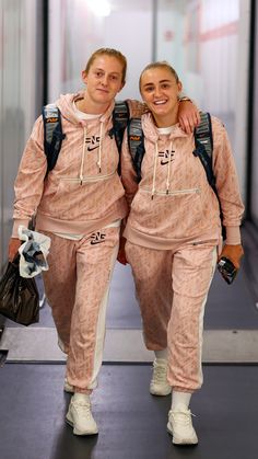 two women in matching pink tracksuits walking down a hallway together with backpacks on their shoulders