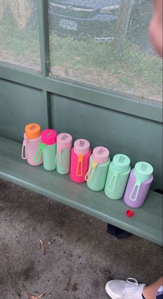 several different colored cups are sitting on a green bench next to a person's feet