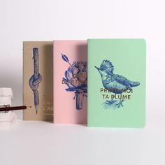 three notebooks are lined up next to each other on a white surface, one has a bird and the other has flowers