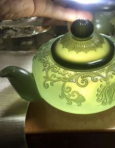 a green teapot sitting on top of a wooden table next to a person's hand