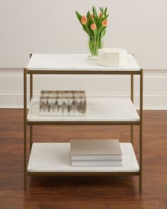 a white table with three shelves holding books and flowers on it, in front of a wall