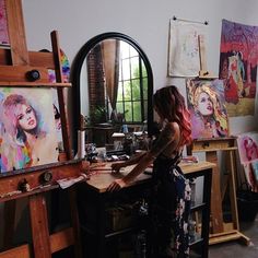 a woman sitting at a desk in front of an easel with paintings on it