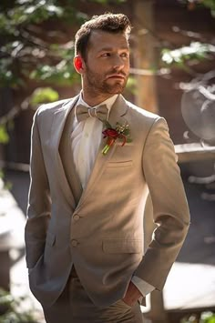 a man wearing a suit and bow tie standing in front of some trees with his hands on his hips