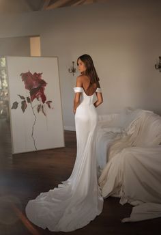 a woman in a white dress is standing near a bed and looking at the wall