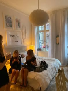 three women are sitting on a bed in a room with white walls and drapes